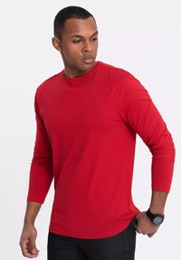 Punainen pitkähihainen puuvillapaita. Pyöreä kaula-aukko, istuva malli, sileä rakenne ja hienovaraiset ommeltiedot. Yhdistetty mustiin farkkuihin.