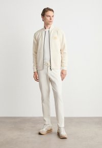 Young man wearing beige zip-up jacket, matching drawstring pants, white shirt, and beige sneakers standing on a concrete floor against a white wall.