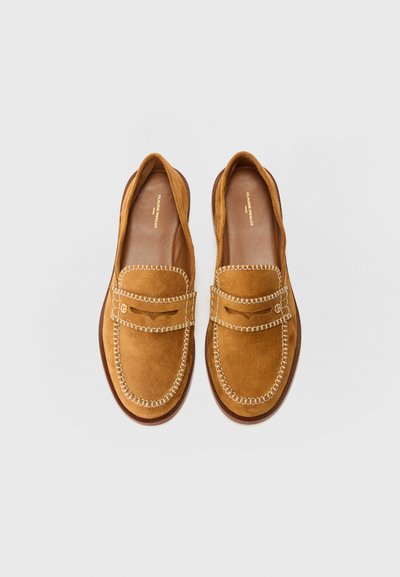 Mocasines de ante marrón con detalle de costura, puntera redondeada y suela de goma texturizada. Interior suave y logotipo de la marca visible.