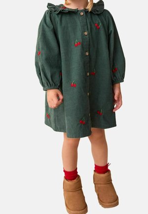 Enfant portant une robe en velours côtelé verte brodée de cerises, avec des boutons en bois, des chaussettes rouges et des bottes d'hiver beige, debout devant un fond uni.
