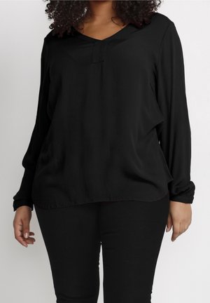 Persoon met krullend haar draagt een losse, zwart blouse met lange mouwen en een V-hals met inkeping, en strak zittende zwarte broek, staand tegen een effen achtergrond.