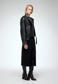 Sort lædermotorcykeljakke med lynlåse, sidebælter og en cropped design, parret med en lang sort kjole og højhalsede sorte støvler.