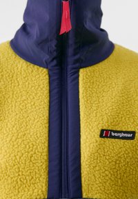 Geel fleecejack met marineblauwe accenten, voorzien van een hoge kraag, ritssluiting en een merklogo. Gelaagd oppervlak voegt diepte toe.