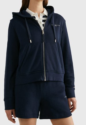 Kobieta ubrana w granatową bluzę z zamkiem i pasujące szorty, na białej koszuli w biało-granatowe paski, stojąca na jednolitym tle.