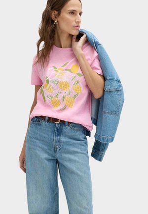 Donna che indossa una maglietta rosa con un motivo di frutta gialla, jeans azzurri chiari, tiene una giacca di jeans sulla spalla, sfondo neutro.