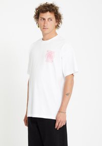 White cotton t-shirt with short sleeves, featuring a pink spiral logo on the left chest. Worn with black pants, model has curly hair.