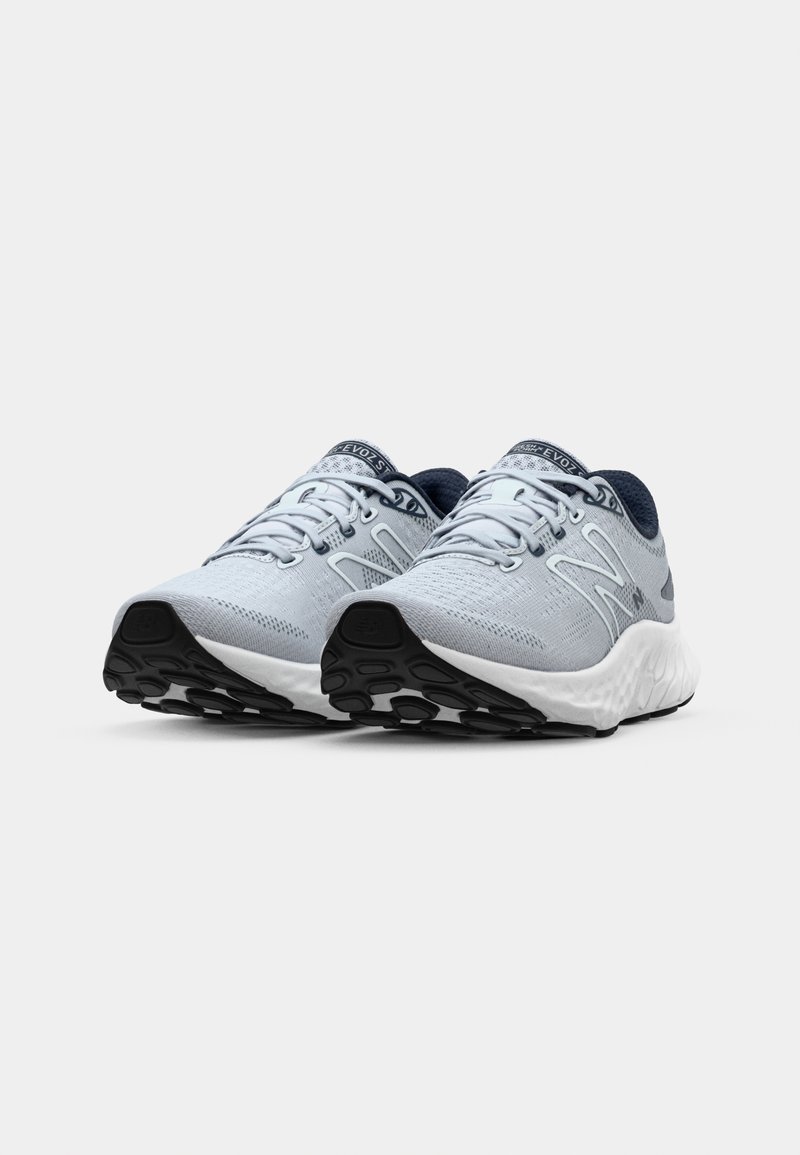 Zapatillas deportivas de color gris claro con parte superior de malla, acentos azules oscuros y una suela blanca texturizada que presenta patrones de tracción negros redondeados.