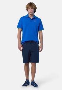 Polo blu con colletto verde, abbinato a pantaloni corti blu scuro. Sneakers sportive completano il look. Design moderno e vestibilità casual.