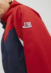 Punainen ja laivastonsininen takki, jossa on teksturoitu pinta, kontrastivalkoiset yksityiskohdat ja pieni brodeerattu Jack & Jones -logo olkapäässä.
