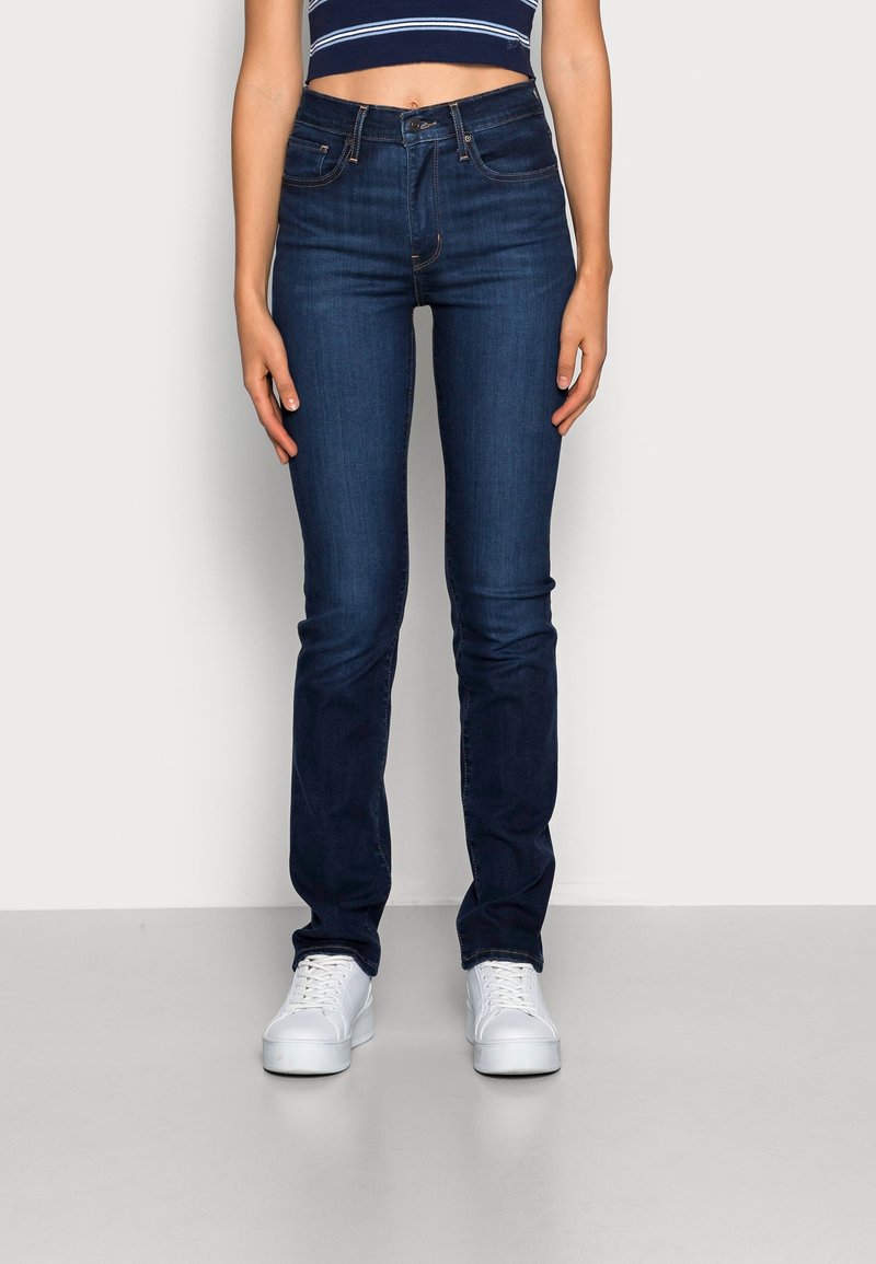 Femme portant un jean slim bleu foncé, des baskets blanches et un crop top bleu marine avec des rayures blanches, debout devant un fond uni.