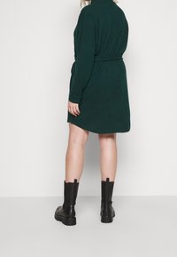 Abito verde scuro a maniche lunghe con vita avvolta da una cintura e texture morbida, abbinato a stivali neri al ginocchio con suola spessa.