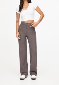 Pantalons larges gris en tissu lisse. Ils présentent une taille haute avec une fermeture à bouton et deux poches latérales. Portés avec un crop top blanc.