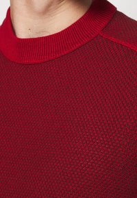 Roter Strickpullover mit strukturiertem Muster, geripptem Halsausschnitt und nahtloser Schulterpartie. Der Stoff wirkt weich und leicht dehnbar.