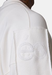 Gros plan sur une personne portant un vêtement blanc avec une pièce texturée présentant des lettres 'é e' en relief et brodées sur le bras supérieur.