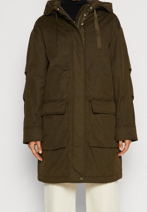 Personne portant un parka à capuche vert olive avec fermeture éclair avant, boutons-pression, grandes poches à rabat et cordons de serrage ajustables à la capuche, associée à un pantalon blanc.