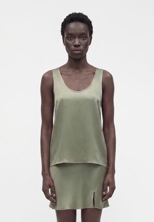 Femme portant un haut en satin vert clair sans manches et une jupe assortie avec une petite fente devant, debout sur un fond blanc uni.