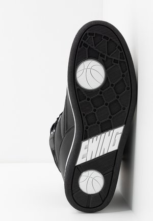 Czarna podeszwa sneakers z białymi ikonami koszykówki i dużym napisem „EWING” wytłoczonym na bieżniku, ustawiona pod kątem na białej powierzchni.
