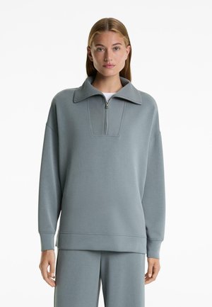 Žena nosi neutralni plavkasto-sivi oversized duksericu s četvrtim zatvaračem i širokim kragnom, u kombinaciji s odgovarajućim hlačama, stojeći na bijeloj pozadini.
