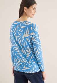 Femme portant un haut à manches longues bleu avec des motifs abstraits de feuilles beiges, assorti à un jean bleu foncé, debout devant un fond uni.