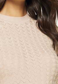 Maglione lavorato a maglia beige con un motivo a onde, scollatura a coste e tessuto texture. Inquadratura ravvicinata che evidenzia i dettagli e la texture.