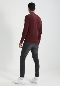 Sudadera burdeos de cuello redondo con puños y dobladillo acanalados, combinada con jeans negros ajustados y zapatillas blancas, vista desde atrás.