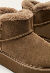 Brune ruskindsankelstøvler med en blød pelsforing og synlige syningsdetaljer, samt en tykk platformssål for ekstra højde.