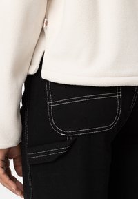 Giacca in pile crema con spacco laterale, abbinata a pantaloni neri con cuciture bianche e una grande tasca sulla coscia destra.