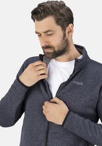 Mann mit Bart, der eine dunkelblaue strukturierte Zip-Jacke über einem weißen Hemd against einem einfachen hellen Hintergrund zippers.