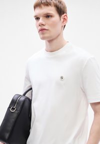 Biele bavlnené tričko s okrúhlym výstrihom a malým logom na hrudi, držené s čiernou koženou taškou s zaoblenými hranami.