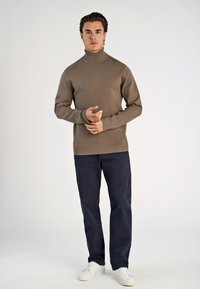 Maglione a collo alto marrone, design aderente. Indossato con pantaloni blu scuro e sneaker bianchi. Modella in piedi con le mani unite, sfondo neutro.