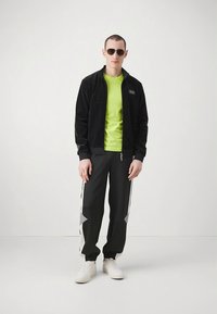 Juoda su užtrauktuku striukė, neoninės žalios spalvos marškinėliai, juodos sportinės kelnės su pilkais akcentais ir balti sportbačiai; modelis dėvi akinius nuo saulės.