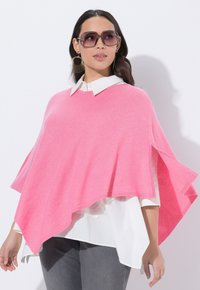 Jasně růžový pletený pončo se širokým výstřihem a asymetrickým lemem přes bílou košili s límečkem. Má dlouhé rukávy a měkkou texturu.