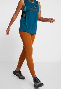 Top senza maniche di colore teal con la scritta "JUST DO IT." stampata in nero, abbinato a leggings arancioni fitted. Scarpe sportive nere con superficie testurizzata.