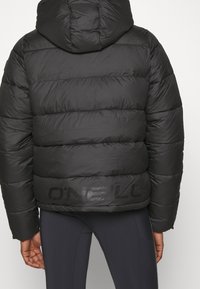 Chaqueta de plumas negra con capucha, que presenta secciones acolchadas y un acabado mate. Logo impreso en negro en la parte baja de la espalda, ajuste ceñido en la cintura.
