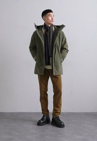 Giacca verde oliva con colletto alto, indossata sopra un maglione beige. Pantaloni in corduroy marroni e scarpe nere chunky. Sciarpa a righe multicolore.