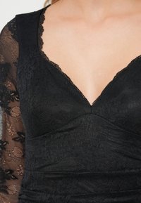 Zwarte kanten jurk met een diepe V-hals, bloemkanten textuur op de mouwen en een getailleerd lijfje met geplooid stofdetail.