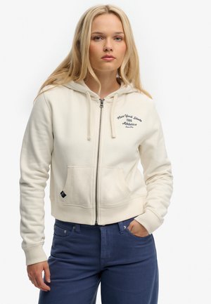 Ung kvinde med langt blondt hår, iført en cremefarvet hættetrøje med lynlås med teksten "New York State 1986 Athletics" og blå bukser, poserer med den ene hånd i lommen.