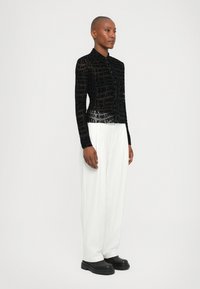 Black sheer top featuring repeating text pattern, high collar, and long sleeves. Paired with white pleated wide-leg trousers and black chunky boots.