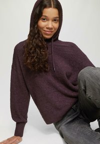 Lila Kapuzenpullover aus strukturiertem Stoff, mit lockerer Passform und gerippten Bündchen, kombiniert mit grauen Jeans.