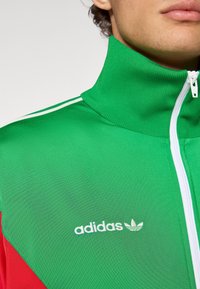 Πράσινο τζάκετ με φερμουάρ και κόκκινες λεπτομέρειες, με ψηλό γιακά και λευκό λογότυπο Adidas. Κατασκευασμένο από ελαφρύ ύφασμα με υφή.