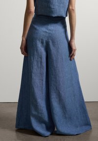 Vista trasera de una persona que lleva pantalones de lino azules de cintura alta y pierna ancha y una camiseta sin mangas a juego, de pie frente a un fondo liso.