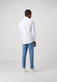 Weißes Langarmhemd mit Kragen, kombiniert mit hellblauen Hosen und weißen Turnschuhen. Glattes Material und taillierte Passform.