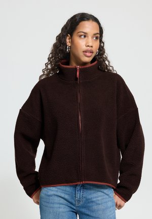 Femme aux cheveux bouclés portant une veste polaire marron foncé zippée et un jean bleu, debout les mains sur les hanches, regardant sur le côté.