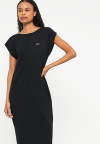 Μαύρο φόρεμα κοντό μανίκι, φτιαγμένο από μαλακό ύφασμα, με χαλαρή εφαρμογή και μια μικρή ετικέτα επωνυμίας στο μπροστινό μέρος. Απαλή υφή και απλός σχεδιασμός.