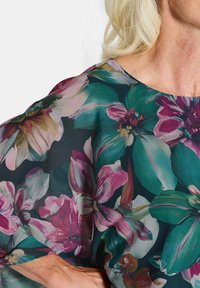 Frau trägt eine dunkelblumige Bluse mit großen pinken, grünen und lila Blumen, die an den Ärmeln und im Brustbereich sichtbar sind.