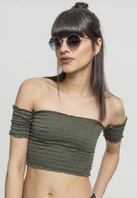 Olivengrønn av- skulder crop top med teksturert smocking, med korte volangermuffser. Modellen har på seg runde solbriller.