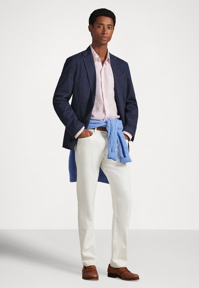 Mladý muž v námorníckom modrom saku, svetloružovej košeli, bielych nohaviciach, hnedých mokasínach a modrom svetri previazanom okolo pásu, stojaci s rukami v kapse.