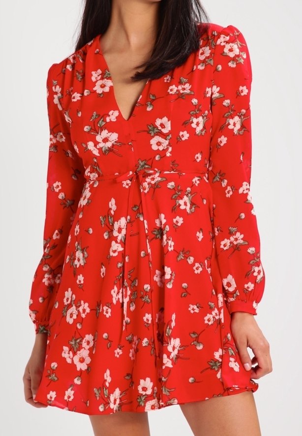 Vestido floral rojo con mangas largas, escote en V y detalle de lazo en la cintura. Presenta una tela ligera con patrones de flores en blanco y rosa.