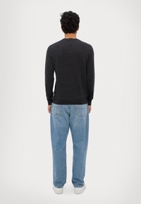 Muž s kudrnatými tmavými vlasy, oblečený v tmavém svetru s dlouhým rukávem, světlých modrých džínách a bílých botách, stojící zády k jasnému pozadí.