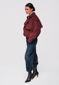 Femme portant une veste courte bordeaux, un jean ample taille haute bleu foncé, et des bottines à talons pointues noires, debout de profil.